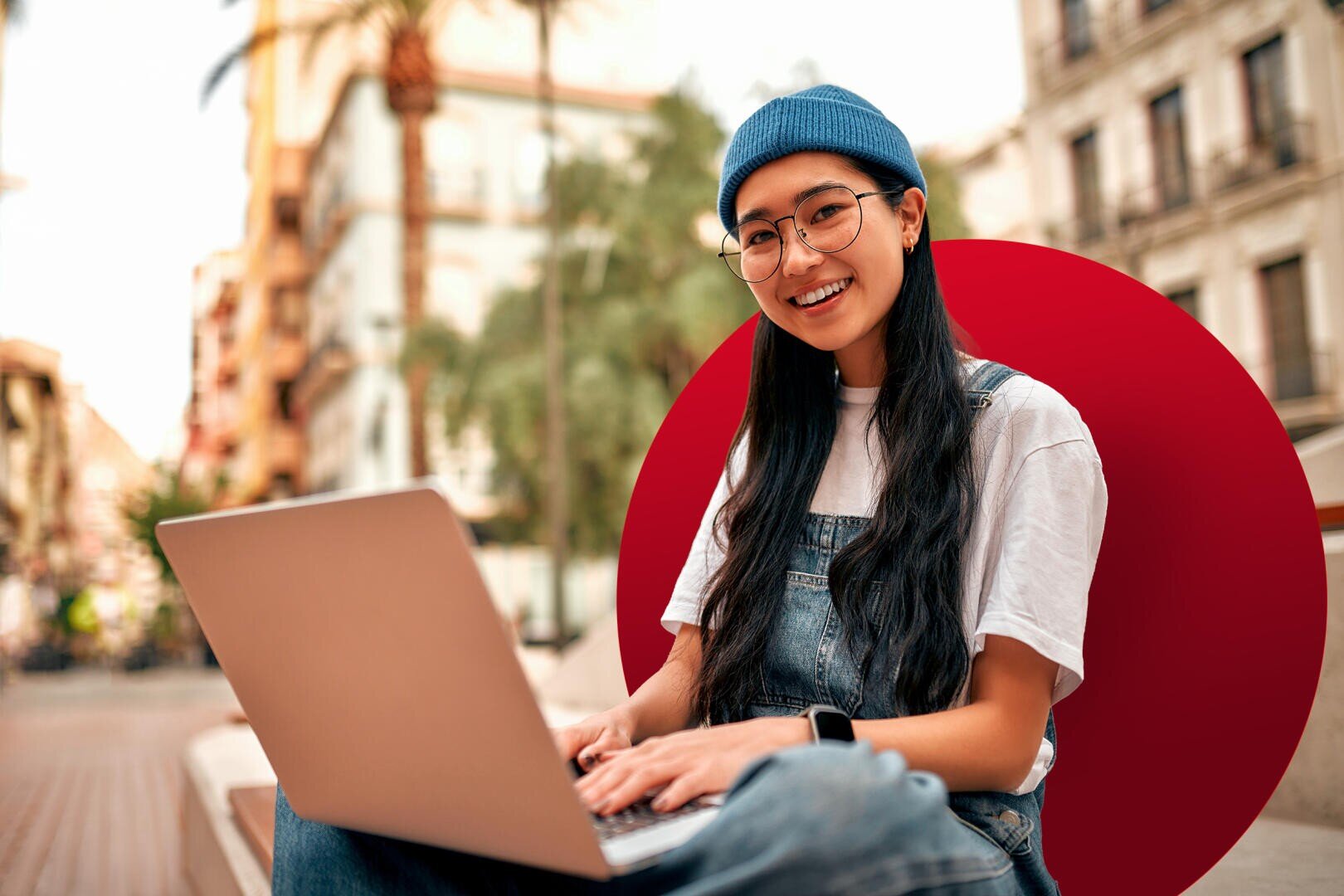 Ein junger Mensch mit langen schwarzen Haaren, Brille und einer blauen Mütze lächelt, während er draußen in einer städtischen Umgebung an einem Laptop arbeitet. Er trägt ein weißes Hemd und eine Jeanslatzhose, im Hintergrund sind Gebäude und Palmen zu sehen.