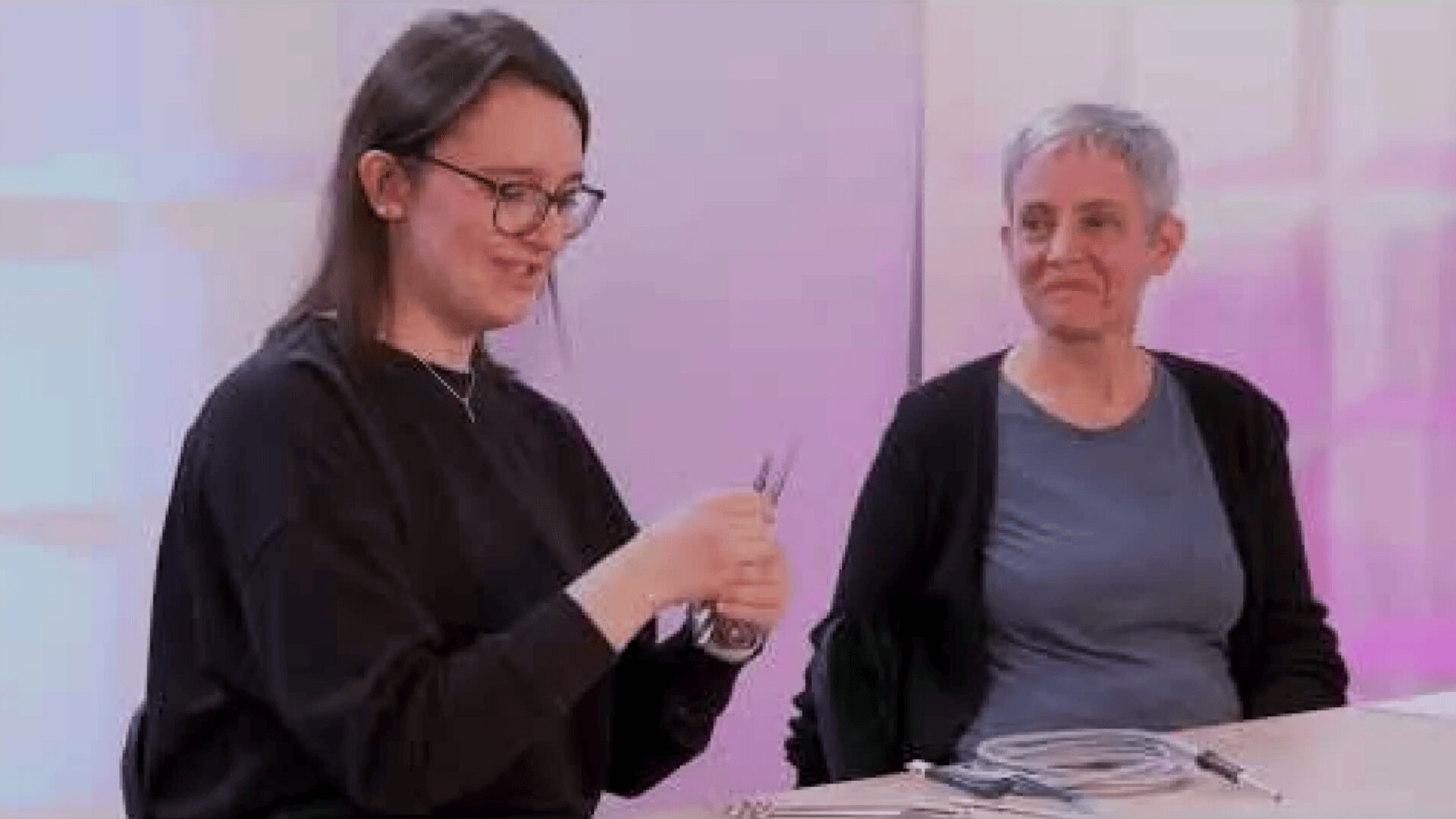 Zwei Frauen sitzen an einem Tisch mit medizinischen Instrumenten. Die eine, mit Brille, hält ein Gerät und lächelt, während die andere, mit kurzen grauen Haaren, sie mit einem warmen Gesichtsausdruck beobachtet. Beide wirken konzentriert und engagiert.