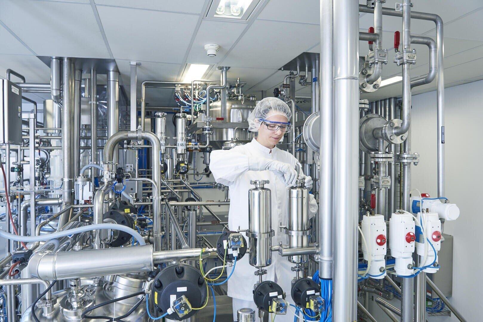 Eine Person in Schutzkleidung und Schutzbrille arbeitet mit Edelstahlgeräten in einem Labor oder einer pharmazeutischen Produktionsstätte, die mit Rohrleitungen und Maschinen ausgestattet ist.