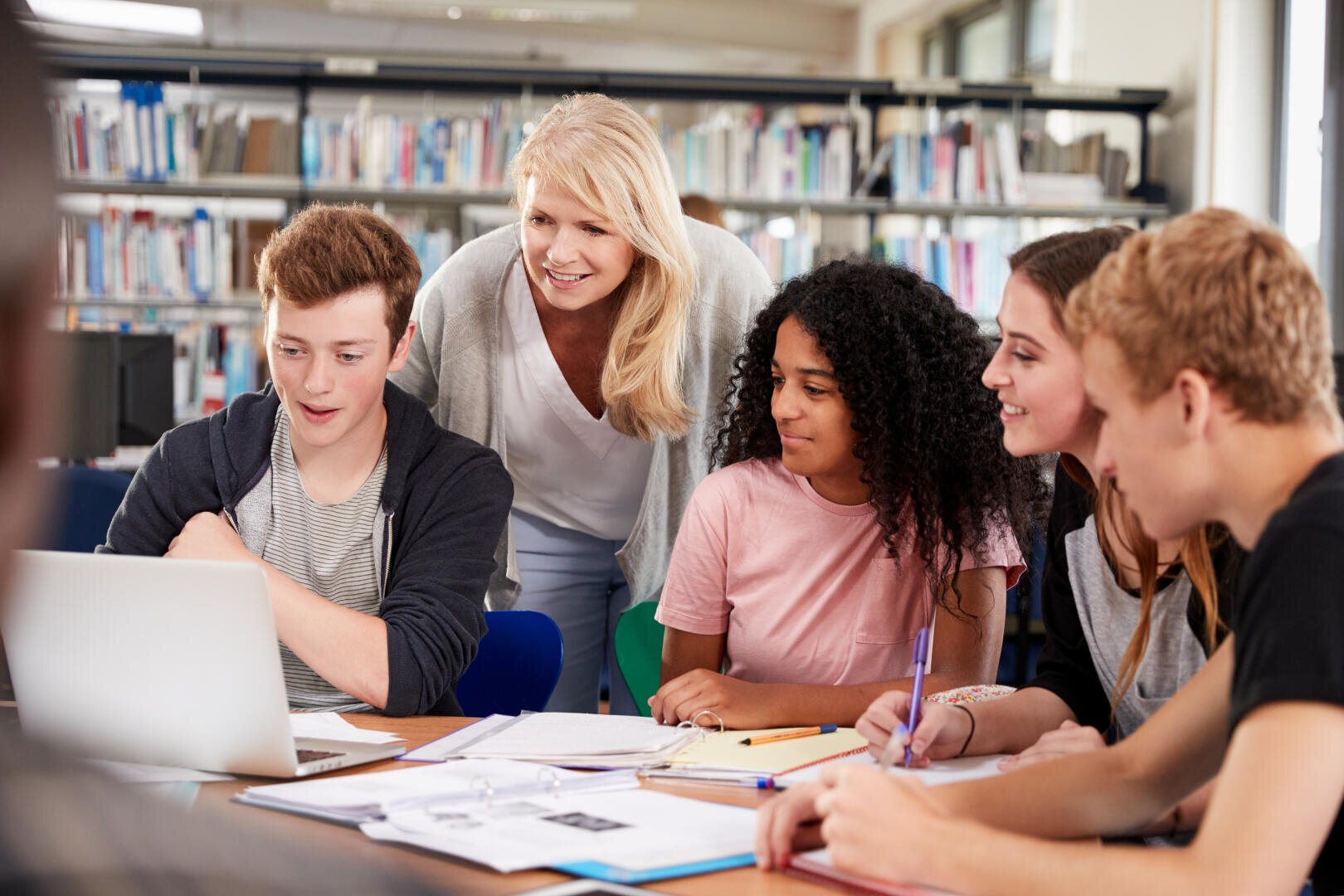 Eine Lehrerin steht lächelnd hinter vier Highschool-Schülern, die sich in einer Bibliothek um einen Tisch mit Notebooks und einem Laptop versammelt haben und in ihre Gruppenarbeit vertieft sind.