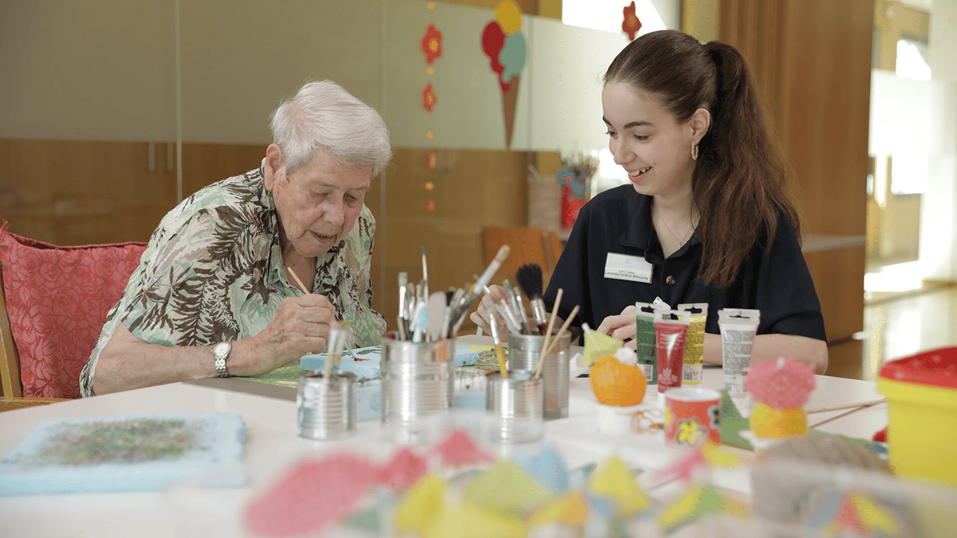 Eine ältere Person und eine junge Frau sitzen an einem mit Malutensilien gedeckten Tisch, malen gemeinsam und lächeln in einem hellen, fröhlichen Raum.