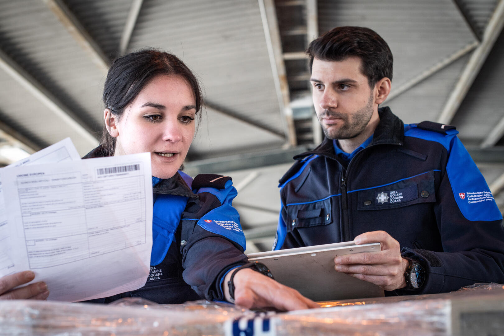 Zwei Zollbeamte in blauen Uniformen untersuchen Papiere und ein Klemmbrett, während sie eine Sendung im Innenbereich unter einem Metalldach kontrollieren.