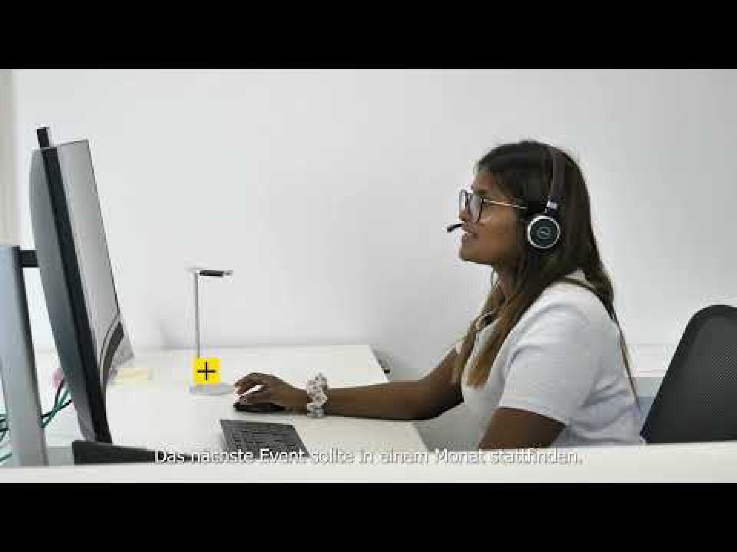 Eine Frau, die eine Brille und ein Headset trägt, sitzt an einem Schreibtisch und schaut lächelnd auf einen Computermonitor in einem hellen Büro. Am unteren Rand des Bildes erscheint ein deutscher Text.