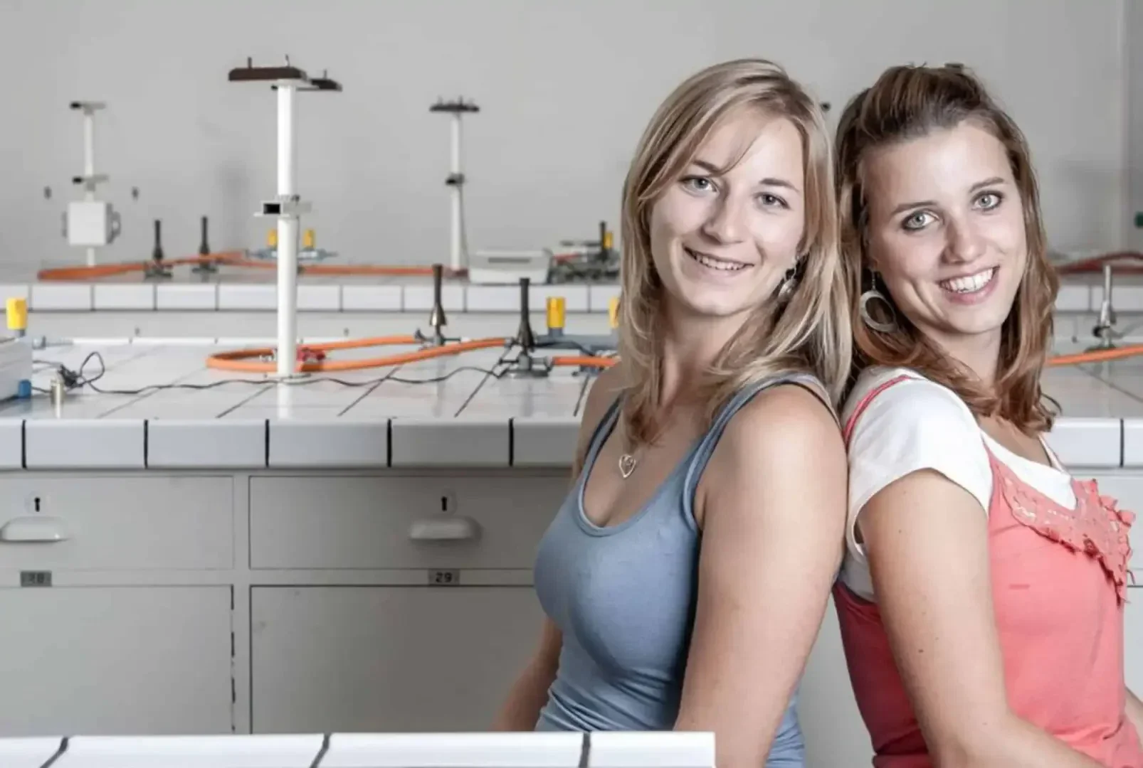 Zwei junge Frauen, die lächelnd Rücken an Rücken in einem wissenschaftlichen Labor sitzen, mit Laborgeräten und weißen Schränken im Hintergrund.