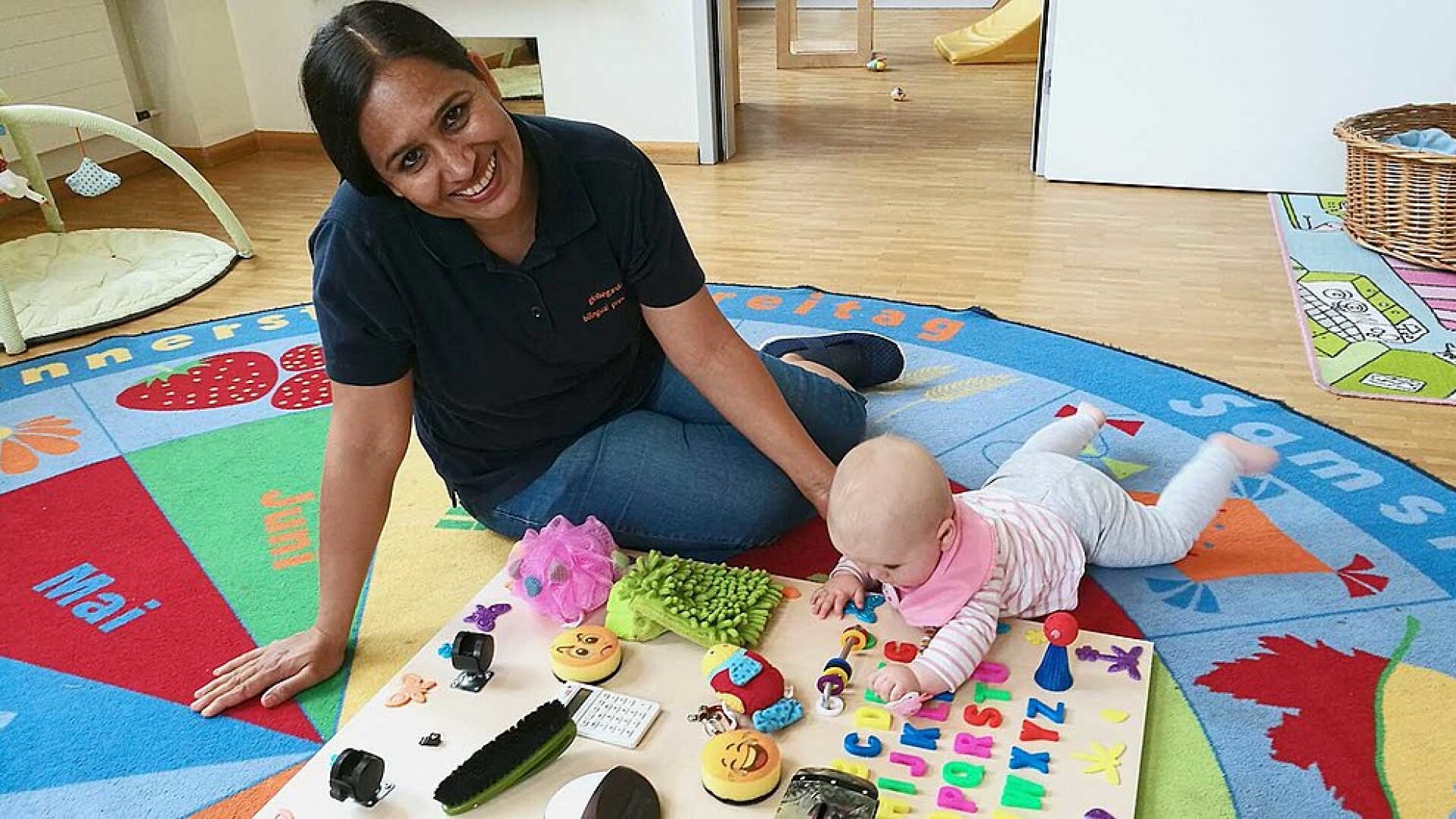 Eine Frau sitzt auf einem bunten Teppich neben einem Baby, das auf dem Bauch liegt und mit verschiedenen Spielzeugen und taktilen Objekten spielt, die auf einem Brett ausgebreitet sind. Beide wirken beschäftigt und glücklich in einem hellen, kinderfreundlichen Raum.