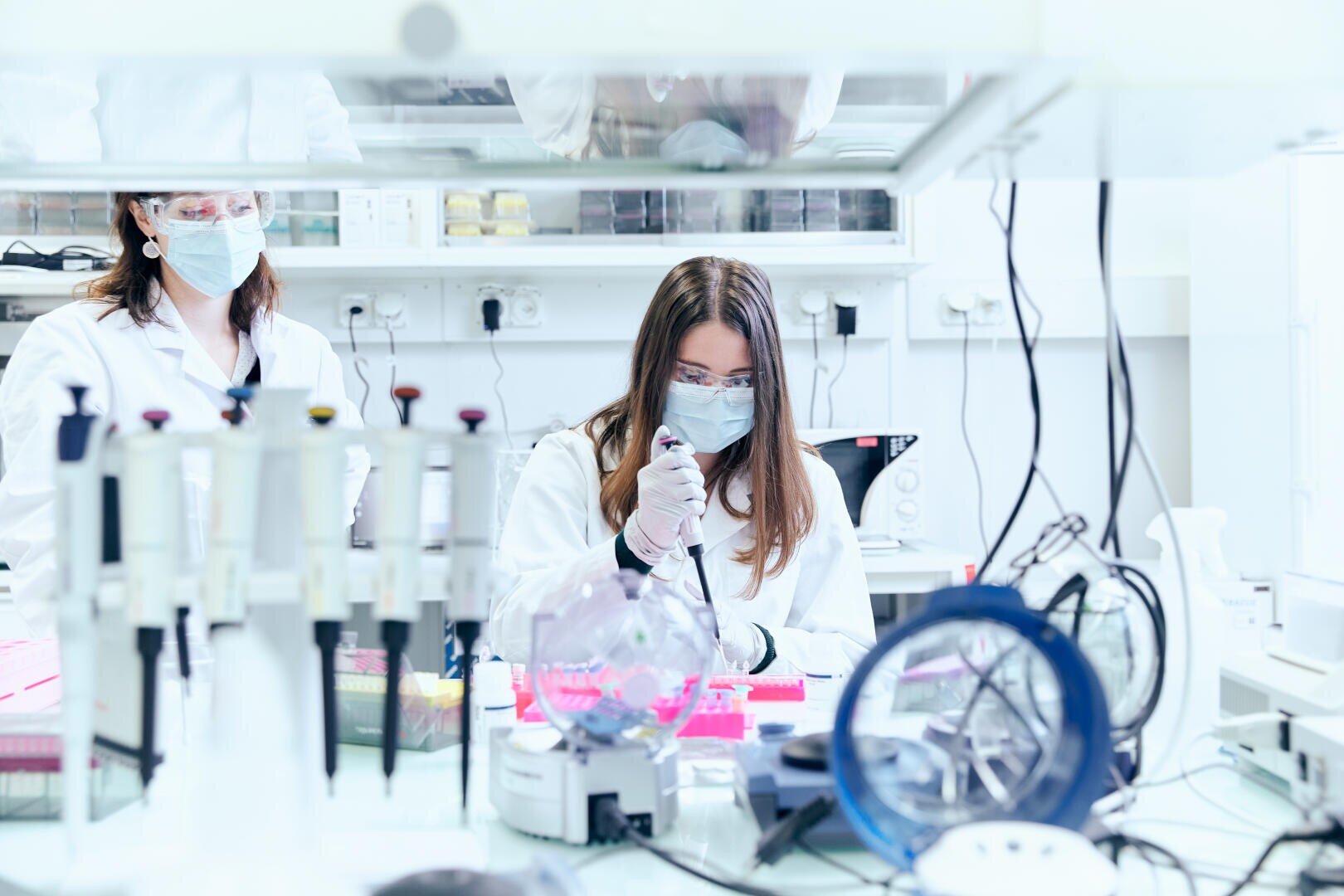 Zwei Wissenschaftler mit Masken und Laborkitteln arbeiten in einem hellen Labor. Einer benutzt eine Pipette mit rosa Proben, während auf dem Tisch im Vordergrund Laborgeräte und Zubehör zu sehen sind.