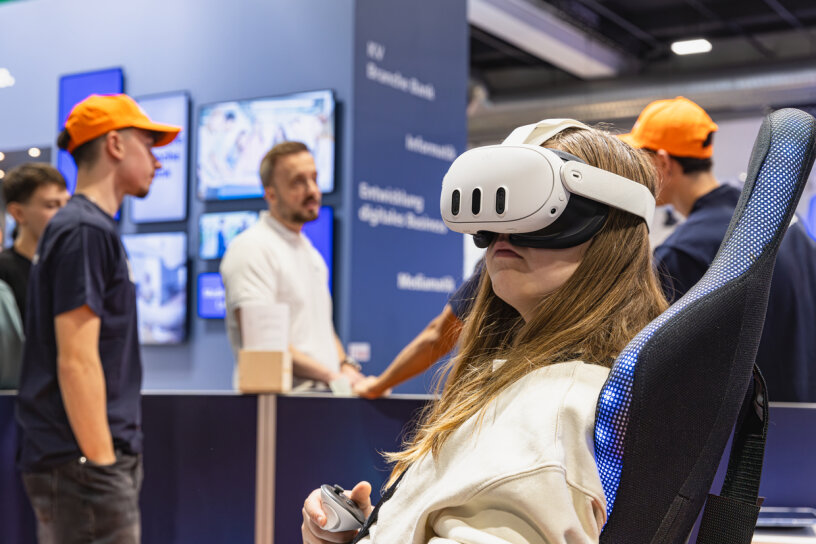Eine Person, die ein weißes Virtual-Reality-Headset trägt und einen Controller in der Hand hält, sitzt auf einem Stuhl bei einer Technologieveranstaltung, während sich im Hintergrund mehrere Personen mit orangefarbenen Hüten unterhalten.
