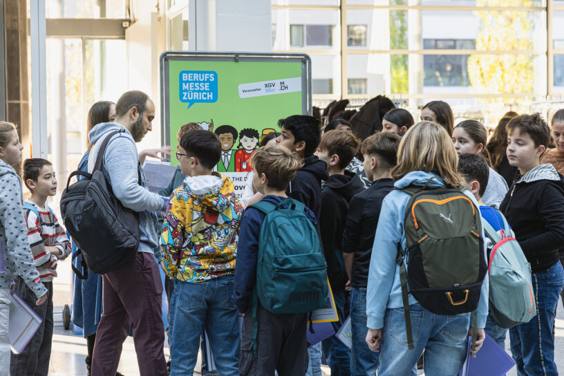 Eine Gruppe von Studenten mit Rucksäcken versammelt sich um einen Mann in der Nähe eines Schildes für die Berufsmesse Zürich in einem hellen Innenraum. Die Schüler wirken aufmerksam und engagiert.