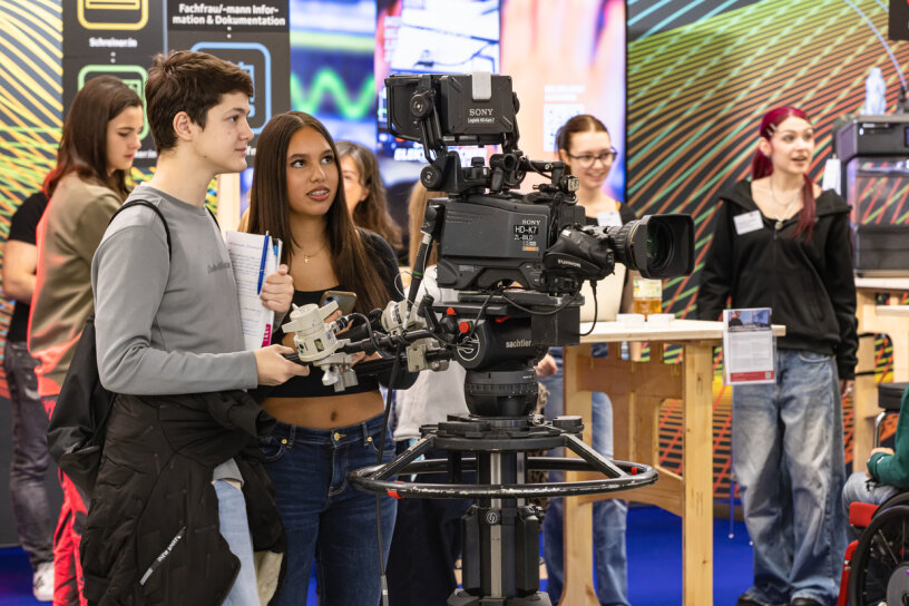 Zwei junge Leute bedienen eine professionelle Videokamera bei einer Indoor-Veranstaltung, während andere im Hintergrund zuschauen und interagieren. Die Umgebung wirkt lebendig und lehrreich, mit bunten Auslagen und Informationspostern.