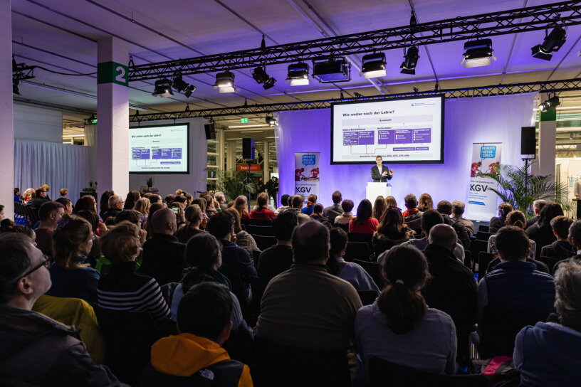 Ein großes Publikum hört einem Redner zu, der auf der Bühne in einem hell erleuchteten Konferenzraum einen Vortrag hält. Zwei große Bildschirme zeigen Informationen hinter dem Redner an. Der Raum ist voll, und die Zuhörer sitzen mit Blick auf die Bühne.