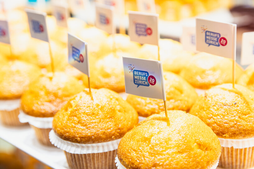 Mehrere goldene Muffins in Pappbechern sind in Reihen angeordnet, auf denen jeweils ein kleines Fähnchen mit der Aufschrift Berufs Messe Zürich und 20 prangt. Das Bild ist hell beleuchtet und hebt die glänzenden Oberseiten der Muffins hervor.
