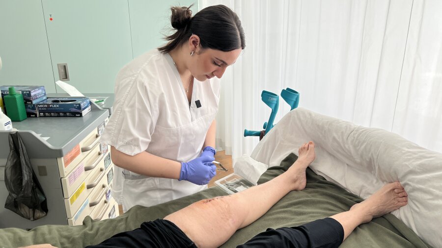 Eine Krankenschwester mit Handschuhen versorgt eine Wunde am Bein eines Patienten, während dieser auf einem Bett in einer medizinischen Einrichtung liegt. Medizinisches Material und Krücken sind in der Nähe zu sehen.