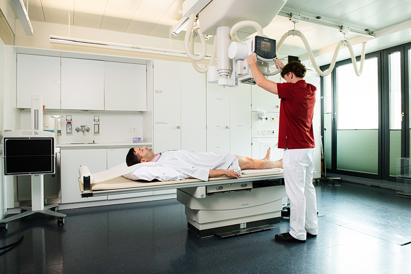 Eine medizinische Fachkraft bedient ein Röntgengerät über einem Patienten, der auf einem Tisch in einem modernen Krankenhauszimmer liegt. Der Patient trägt einen Krankenhauskittel, und der Raum ist mit medizinischen Geräten und Schränken ausgestattet.