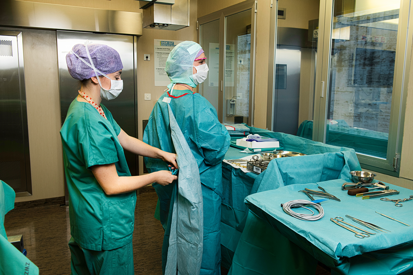Zwei medizinische Fachkräfte in Kitteln und Masken bereiten sich in einem Operationssaal auf einen Eingriff vor. Der eine hilft dem anderen beim Anlegen eines OP-Kittels. Sterile chirurgische Instrumente liegen auf einem Tisch in der Nähe bereit.
