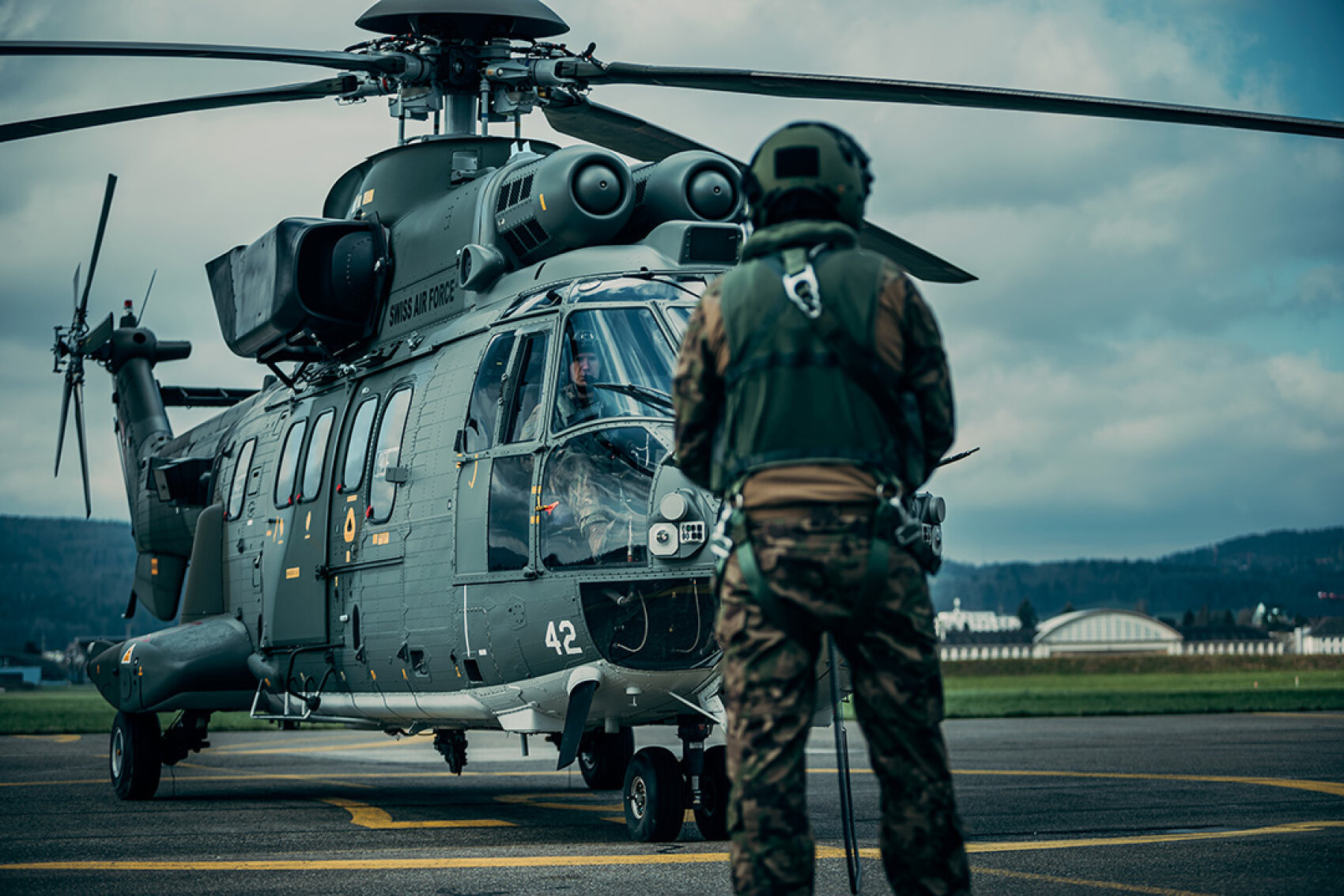 Ein Militärhubschrauber steht mit stillstehenden Rotoren auf einem Rollfeld, während ein uniformierter Soldat mit Helm im Vordergrund steht und auf das Fluggerät blickt. Der Himmel ist bewölkt und im Hintergrund sind Hangars zu sehen.