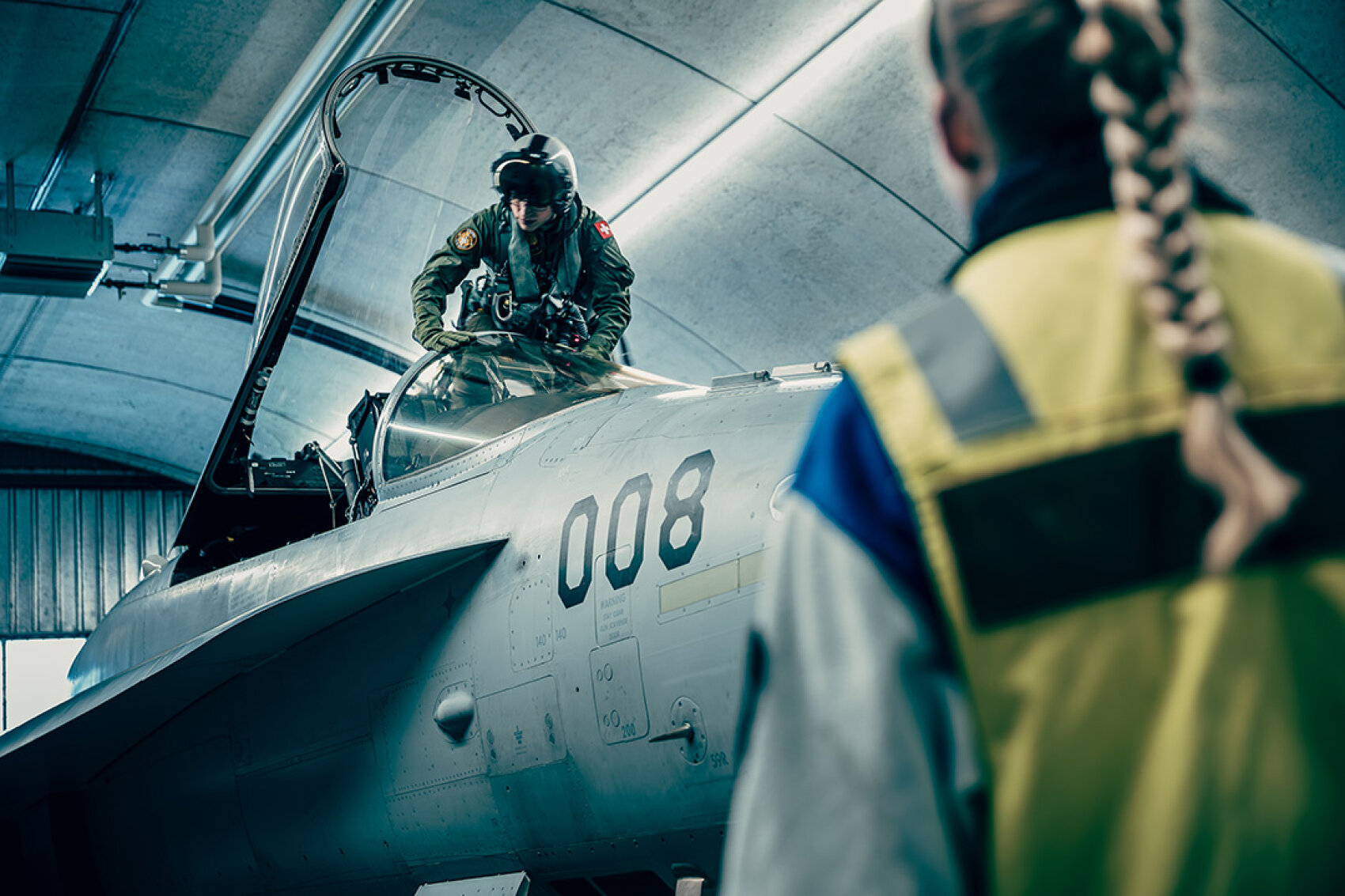 Ein Pilot bereitet sich darauf vor, das Cockpit eines Kampfjets mit der Nummer 008 in einem Hangar zu verlassen, während eine Person in einer gelben Sicherheitsweste mit einem geflochtenen Pferdeschwanz im Vordergrund steht und zuschaut.