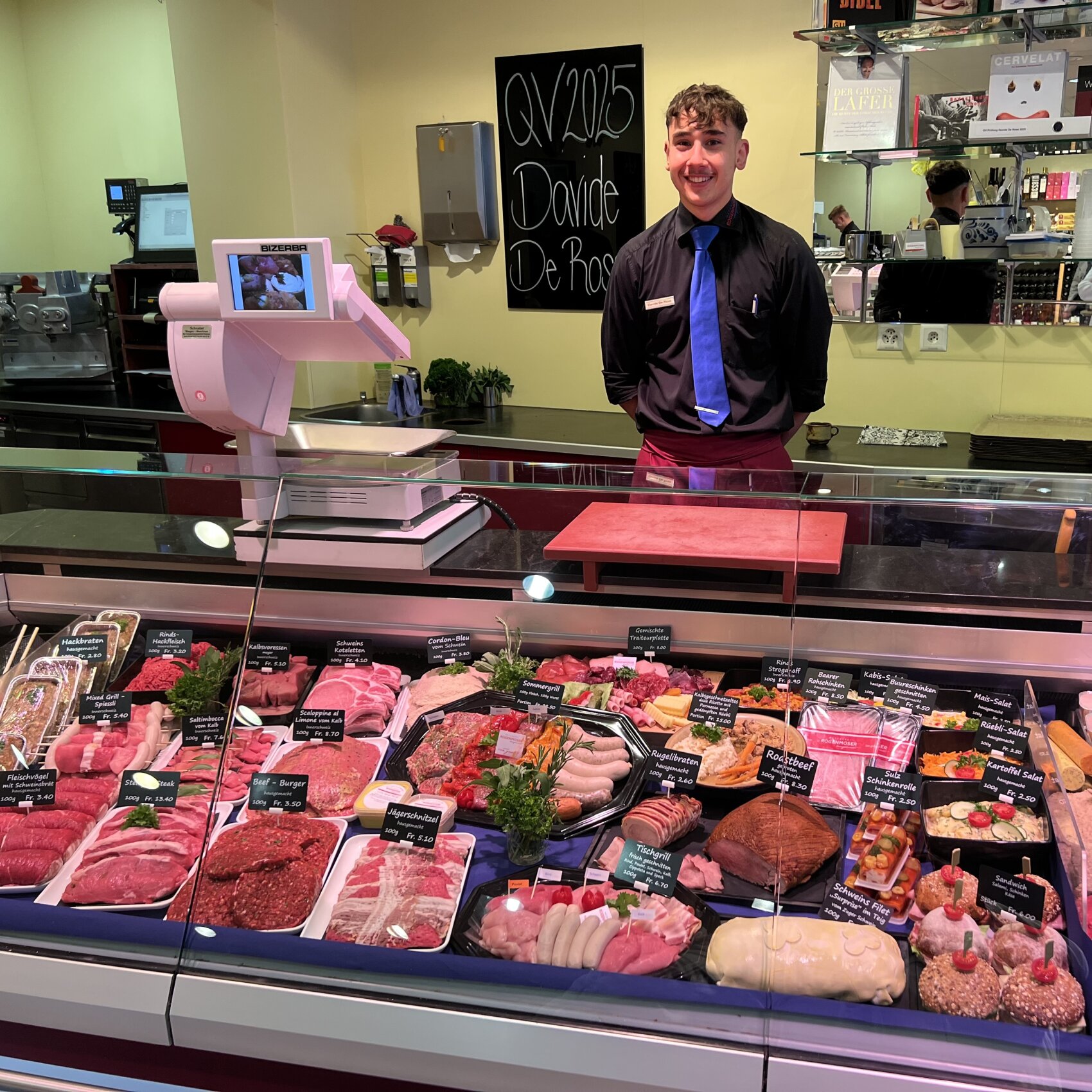 Ein lächelnder Metzger steht in einem hell erleuchteten Geschäft hinter einer Glasvitrine mit verschiedenen Fleisch- und Wurstsorten und Feinkostartikeln. Schilder mit Etiketten und Preisen sind auf den Lebensmitteln zu sehen.